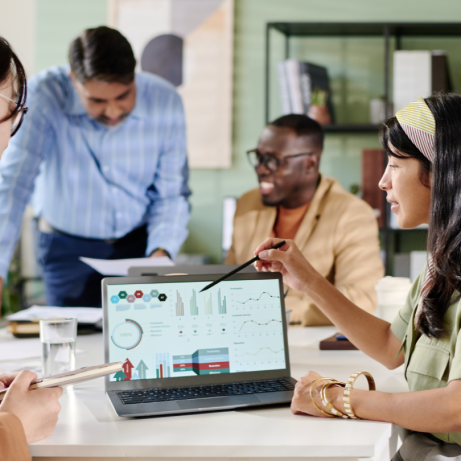 A group of people are gathered around a laptop displaying charts and graphs, with one person pointing at the screen.