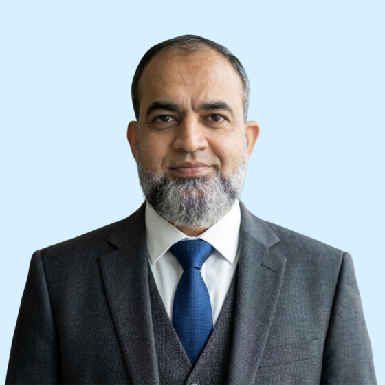 Portrait of a man in a dark suit with a light shirt and blue tie, with a neatly trimmed gray beard, facing the camera against a pale blue background.