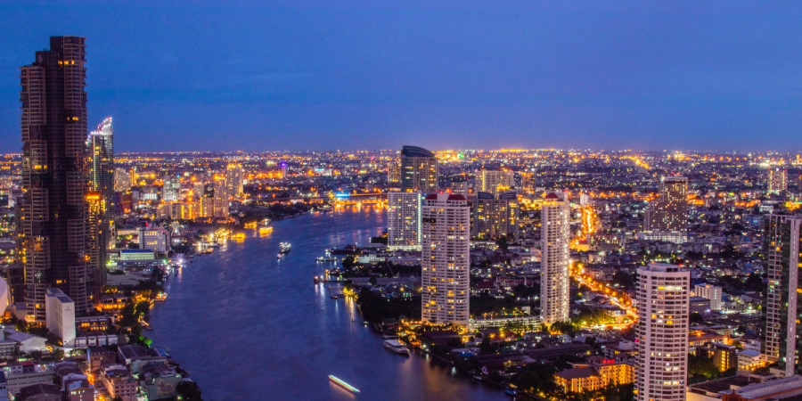 a panoramic night time view of a wide river flowing through a densely populated city with numerous illuminated skyscrapers and buildings along its banks
