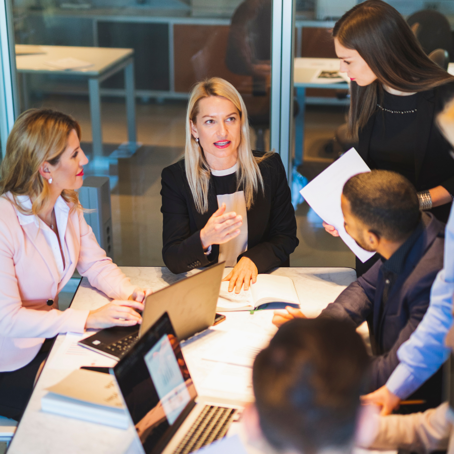 A group of people in a meeting around a table with laptops and papers.