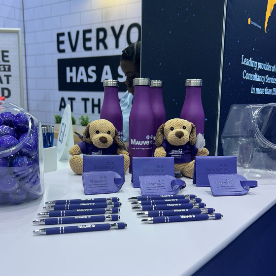 A trade show booth display featuring purple water bottles, two stuffed dogs wearing company branded shirts, purple pens, and a glass jar filled with purple candies.