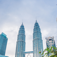 the Petronas Twin Towers are visible among other skyscrapers, with lush green trees in the foreground under a cloudy sky