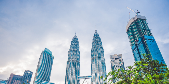 the Petronas Twin Towers are visible among other skyscrapers, with lush green trees in the foreground under a cloudy sky