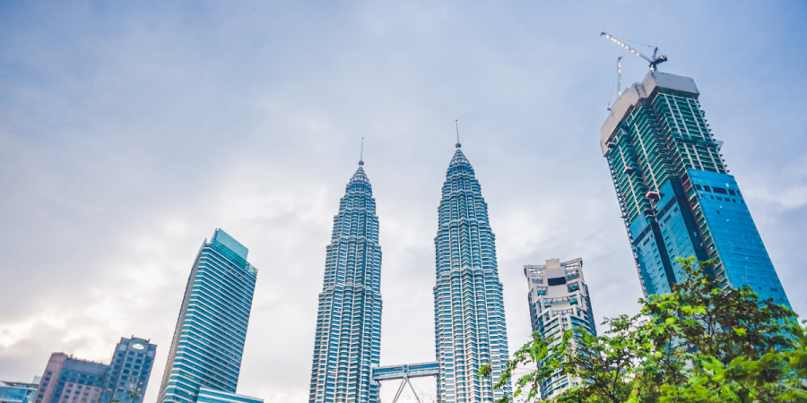 the Petronas Twin Towers are visible among other skyscrapers, with lush green trees in the foreground under a cloudy sky