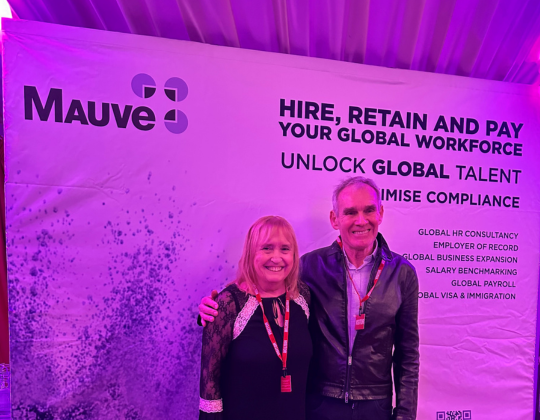 Ann & John Ellis stand in front of a Mauve Group banner that reads "Hire, retain and pay your global workforce", "Unlock global talent", "Optimise compliance", and "Trusted worldwide for 30 yrs".