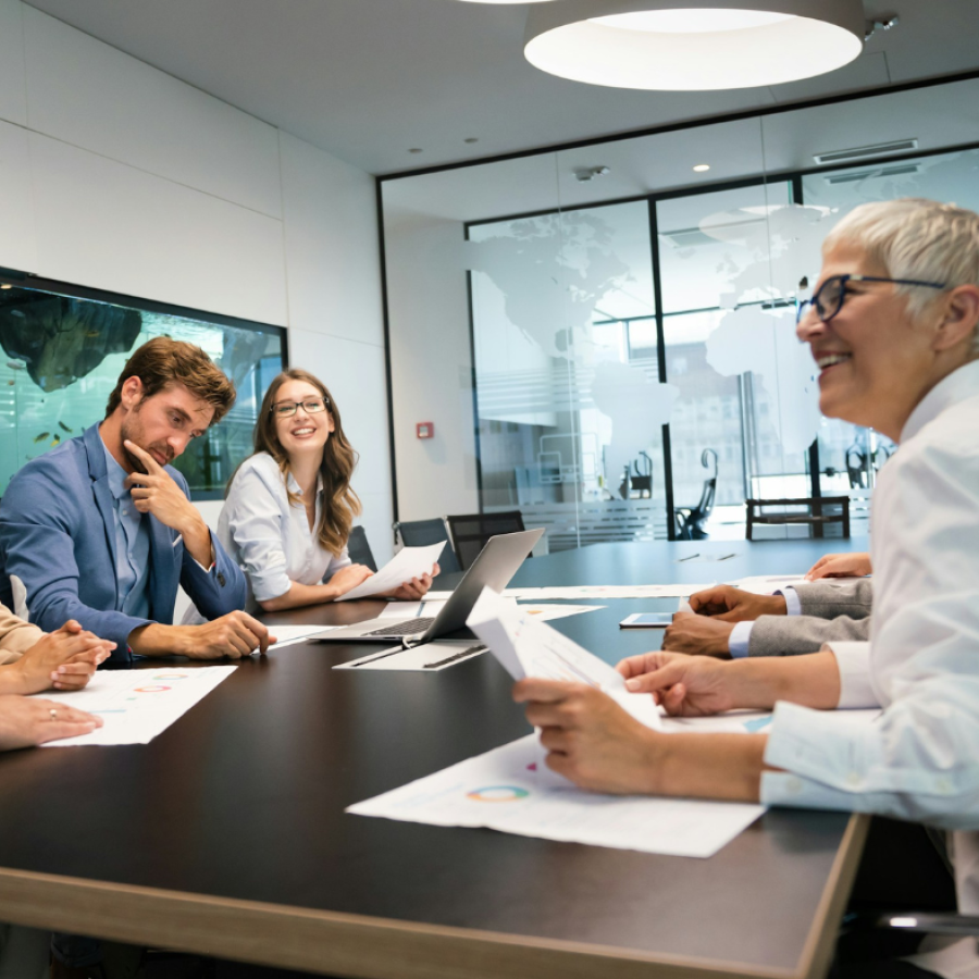 A group of business professionals gathered around a conference table, looking at documents and laptops during a meeting.