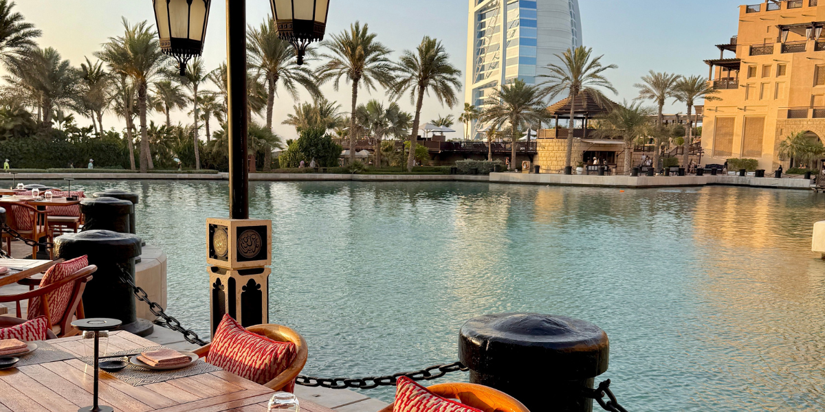 Outdoor dining setup on a wooden deck by calm water, with lanterns and cushions, palm trees, and a tall sail-shaped building across the marina under a clear blue sky.