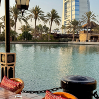 Outdoor dining setup on a wooden deck by calm water, with lanterns and cushions, palm trees, and a tall sail-shaped building across the marina under a clear blue sky.