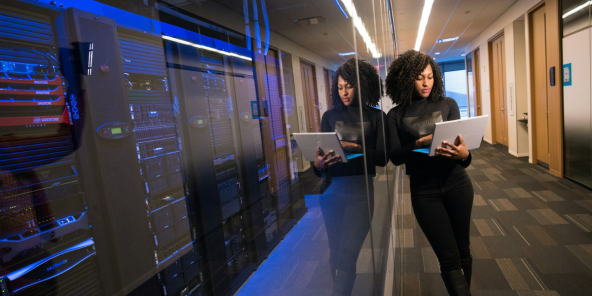 Woman is leaning against a wall of computer servers holding a laptop