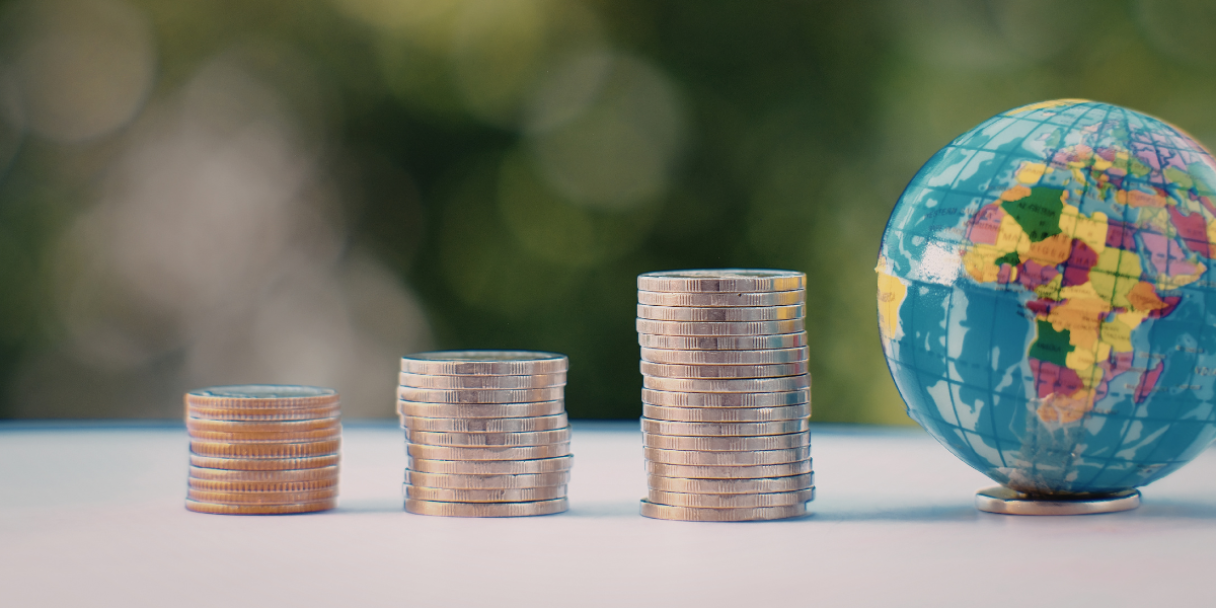 A stack of silver coins forms rising steps beside a blue globe on a table, with a blurred green outdoor background suggesting growth and global finance.