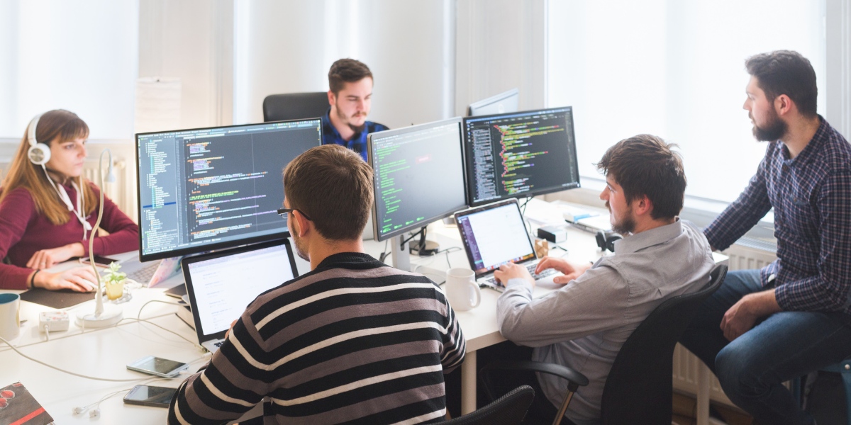 A group of people in a bright office collaborating at a table with multiple monitors showing code, while one person works at a laptop and others discuss the screens.