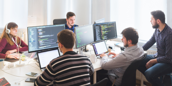 A group of people in a bright office collaborating at a table with multiple monitors showing code, while one person works at a laptop and others discuss the screens.