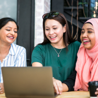 three Asian women are sitting at a table with a laptop and coffee cups, laughing