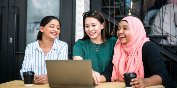 three Asian women are sitting at a table with a laptop and coffee cups, laughing