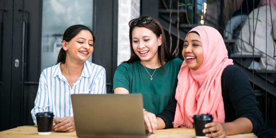three Asian women are sitting at a table with a laptop and coffee cups, laughing